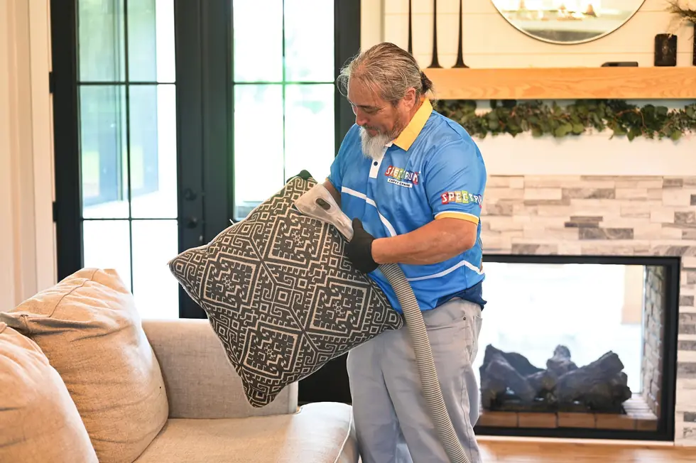 Spectrum technician in a blue polo vacuuming a patterned cushion in a bright living room with a fireplace
